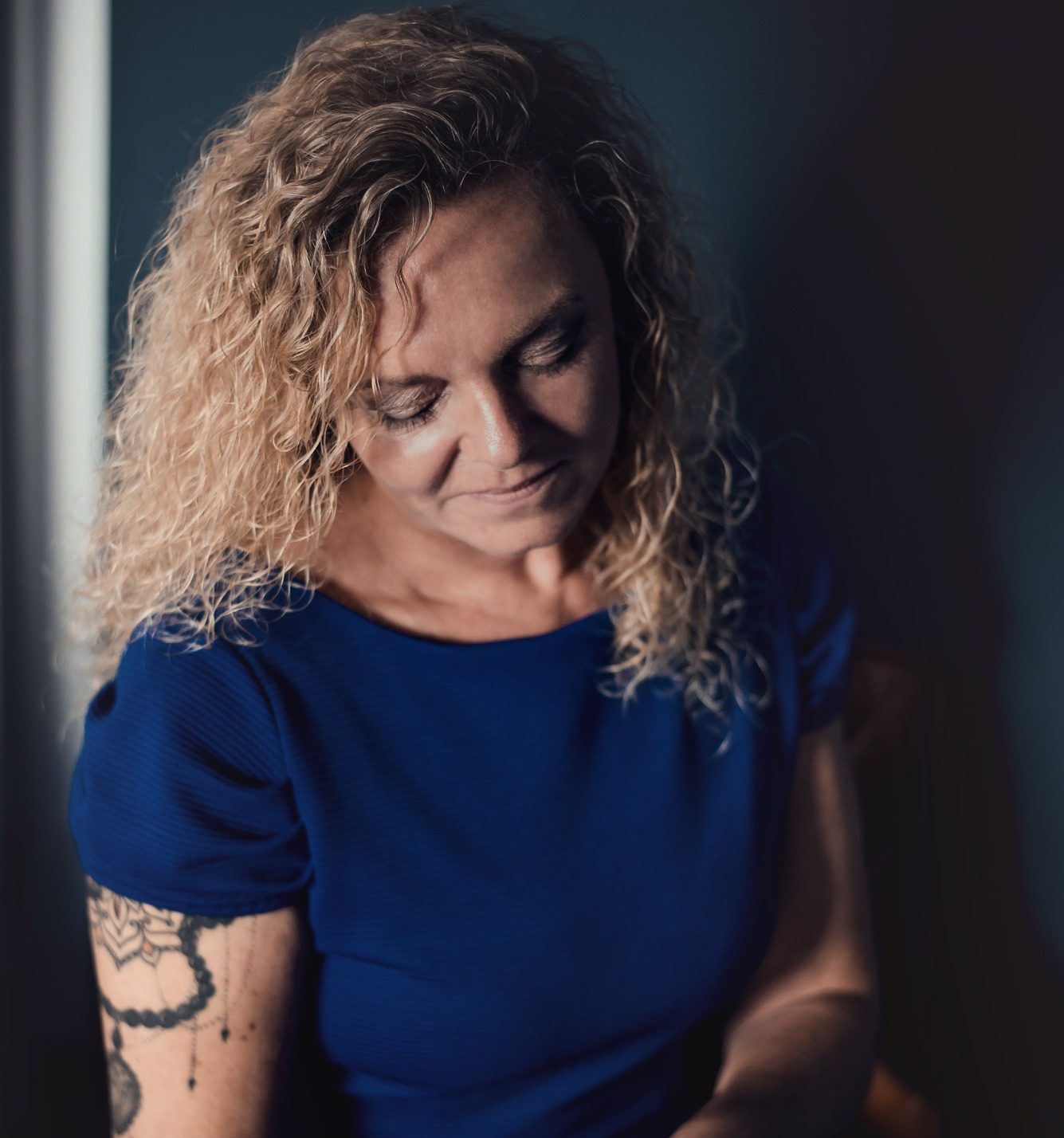 Eine nachdenkliche Frau mit lockigem Haar und Tatoos, die in einem blauen Kleid sitzt.
