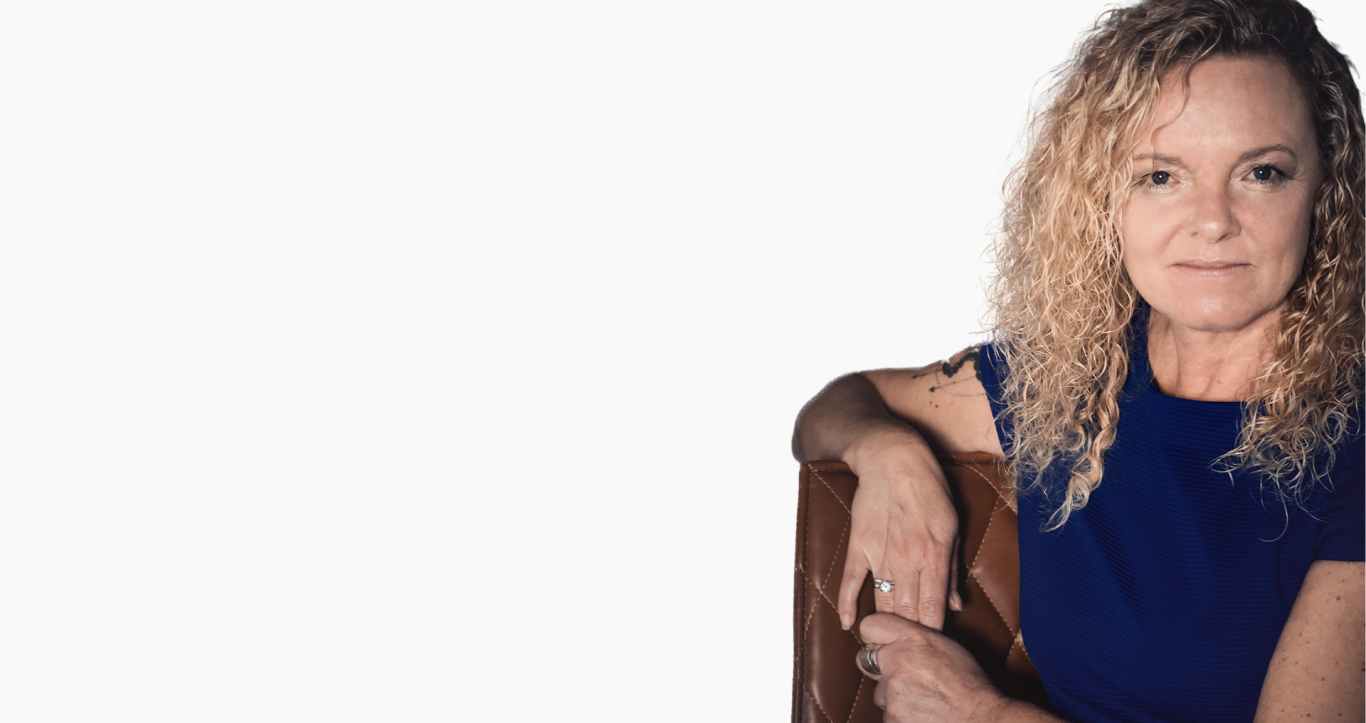 Frau mit lockigem, blondem Haar, in blauer Kleidung, schaut lächelnd in die Kamera.